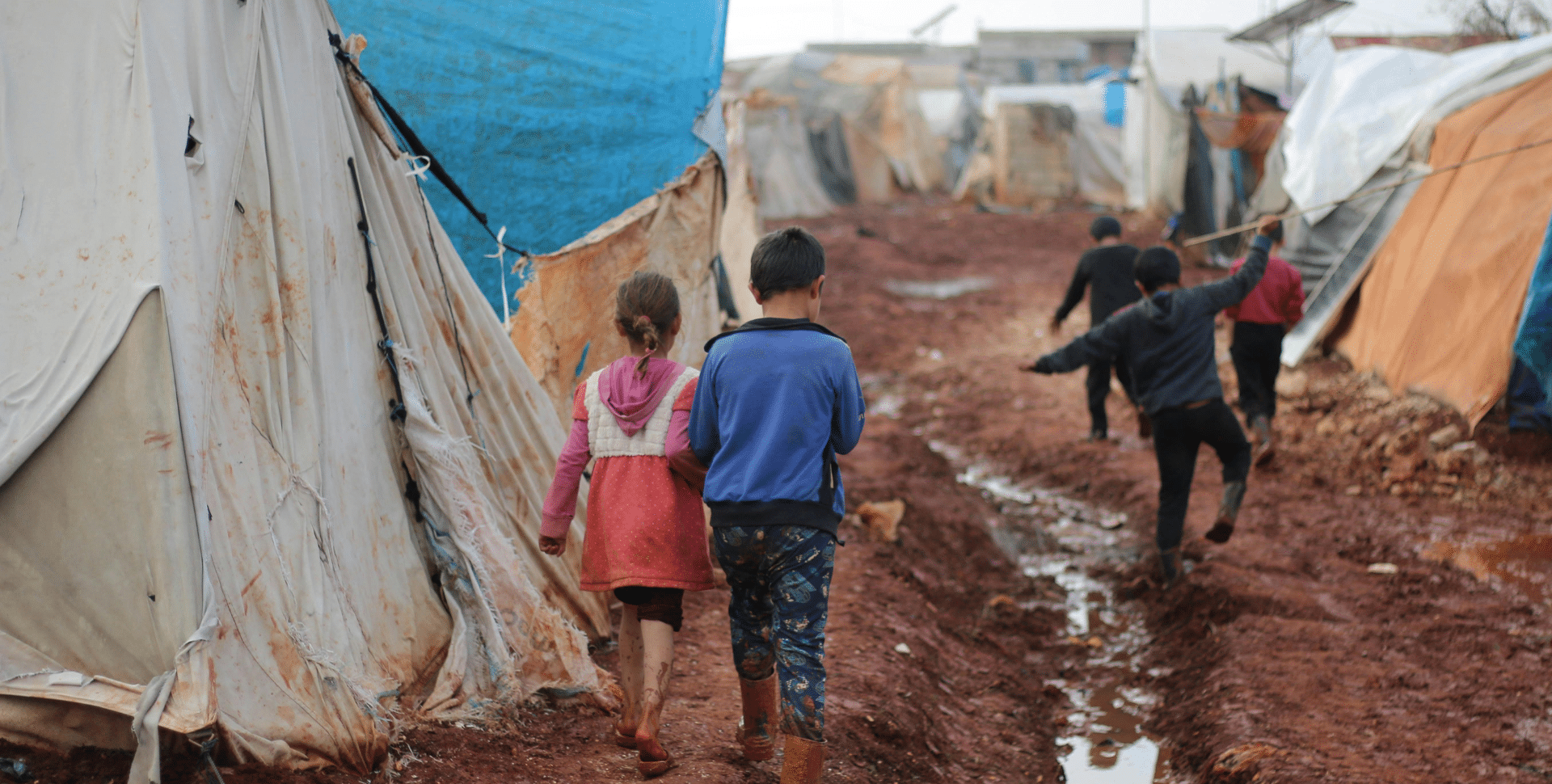 A group of children walk on a muddy path between tents at a refugee camp