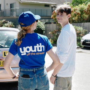 A Youth Off The Streets volunteer stands with a young person who they are helping teach how to drive, through the Youth Off The Streets iDrive Program.